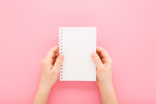 Little child girl hands holding white copybook on light pink table background. Pastel color. Closeup. Empty place for text. Point of view shot. Top down view.
