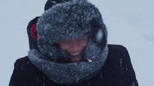 Happy boy in a warm earflap hat throwing snow and laughing. Portrait of a child playing outside during a heavy snowfall