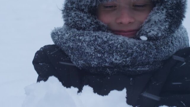 Close-up portrait of a happy boy in a fur ushanka hat covered with frost and snow. The child smiles sincerely while looking at the camera during a heavy winter snowfall.