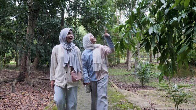 Two young Asian Muslim sisters walking through a lush forest park, chatting and enjoying nature together under the morning sun