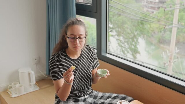 Hotel corner snack by window, guest sampling pastry and tea while seated on bench. Urban greenery and power lines visible through glass. Relaxed candid vibe, soft daylight and informal morning