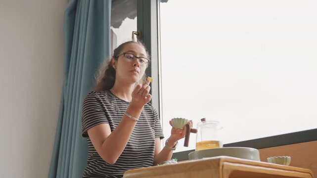 Hotel table tasting with city view, guest bites pastry while sipping tea and lifting eyes toward window light. Bright interior with pendant lamp, tray with teapot and small bowls, upbeat indulgent