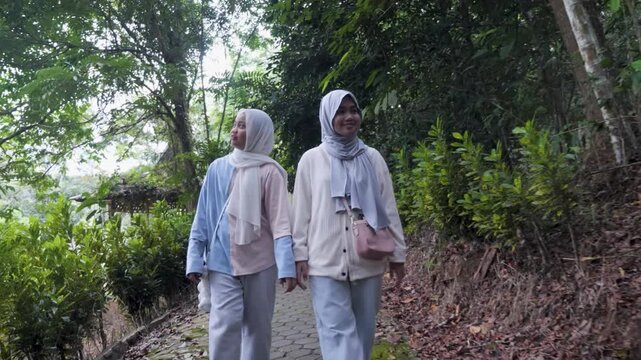 Two young Asian Muslim sisters walking through a lush forest park, chatting and enjoying nature together under the morning sun