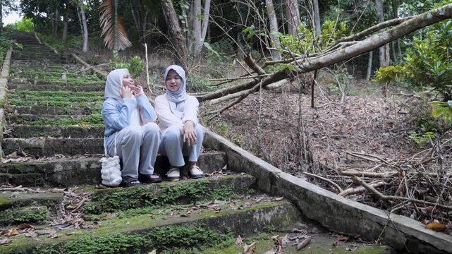 Two young Asian Muslim sisters sitting on forest park stairs, chatting and enjoying nature together under the morning sun
