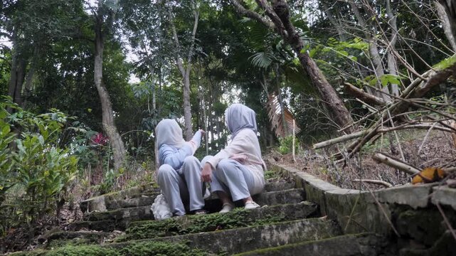 Two young Asian Muslim sisters sitting on forest park stairs, chatting and enjoying nature together under the morning sun