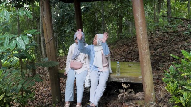 Two young Asian Muslim girls sitting and resting in a wooden gazebo, chatting and enjoying nature together under the morning sun in a forest park