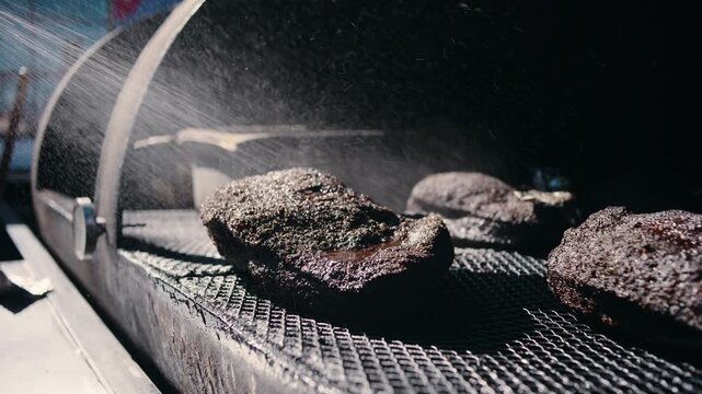 A pitmaster in a canvas apron walks past a large outdoor woodpile carrying a split log of firewood for the smoker in daylight