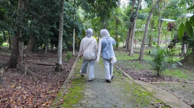 Two young Asian Muslim sisters walking through a lush forest park, chatting and enjoying nature together under the morning sun