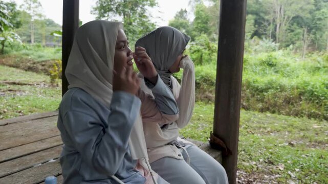 Two young Asian Muslim girls sitting and resting in a wooden gazebo, chatting and enjoying nature together under the morning sun in a forest park
