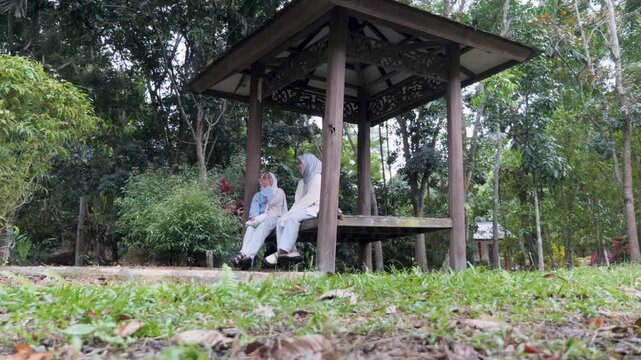 Two young Asian Muslim girls sitting and resting in a wooden gazebo, chatting and enjoying nature together under the morning sun in a forest park
