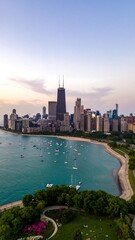 Obraz premium An aerial shot showcases a vibrant city skyline alongside a sparkling bay, with boats and a lush green park at the bottom. The sky blazes with sunset hues