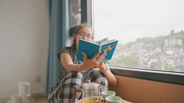 Hotel bay window guest reading guidebook, travel planning gesture, tea service on low table, glasses and pages, harbor hillside view, reflective solo traveler moment, itinerary preparation