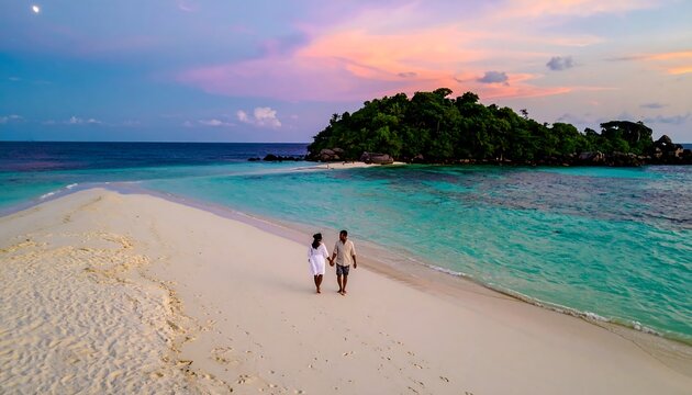 An aerial view of a couple walking hand-in-hand on a sandy isthmus connecting to a lush, tropical island. The sky showcases vibrant colors at sunset