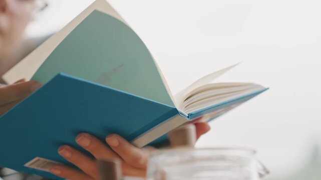 Woman reading blue notebook in hotel, closeup hands turning pages, coffee jar on tabletop, turquoise stickered cover, relaxed guest on sofa, soft natural light and calm leisure atmosphere.