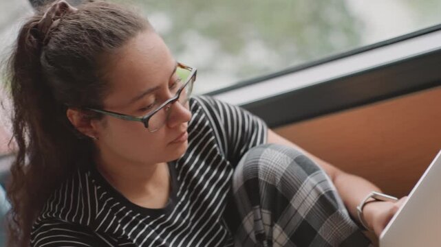 Young woman with laptop hotel window, legs tucked and plaid pants, pensive gaze while checking itinerary and notes, adjusting glasses, soft greenery outside, relaxed lounge mood and quiet