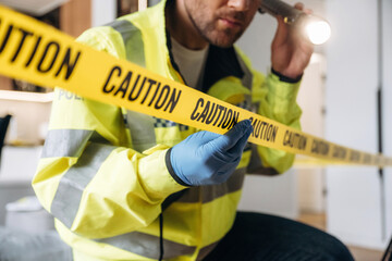 Forensic expert examining caution tape at a crime scene, wearing gloves and a reflective jacket,...