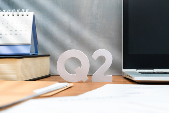 Letters Q2 on a wooden desk alongside a laptop, a book, and a desk calendar. Second quarter business results, fiscal planning, and corporate performance reporting concept.