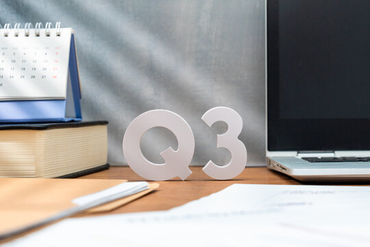 Letters Q3 on a wooden desk alongside a laptop, a book, and a desk calendar. Third quarter business results, fiscal planning, and corporate performance reporting concept.