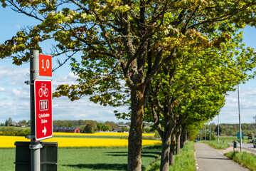 Red bicycle route sign (Cykelleden Sk&aring;ne 101) near rapeseed fields in Swedish countryside, Skane Concept of cycling tourism, eco-friendly transport and outdoor lifestyle.
