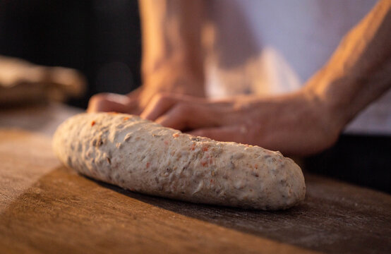 Baker hands shaping long loaf of multi-grain bread dough on rustic wooden table, preparation of food ingredient for baking process, close-up view of culinary activity, artisanal bakery concept