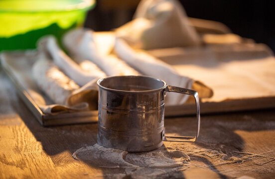 Metal flour sifter stands on wooden table covered with scattered white flour, loaves of unbaked dough rest in soft focus background, preparation for baking bread in home kitchen