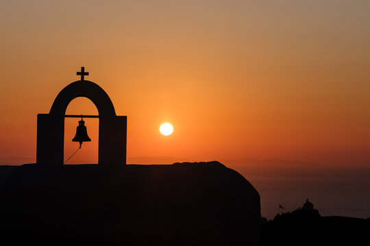 View of a the silhouette of a Greek Orthodox Church, with a bell and a cross at the top and a spectacular orange sunset in the background in Ios cyclades Greece