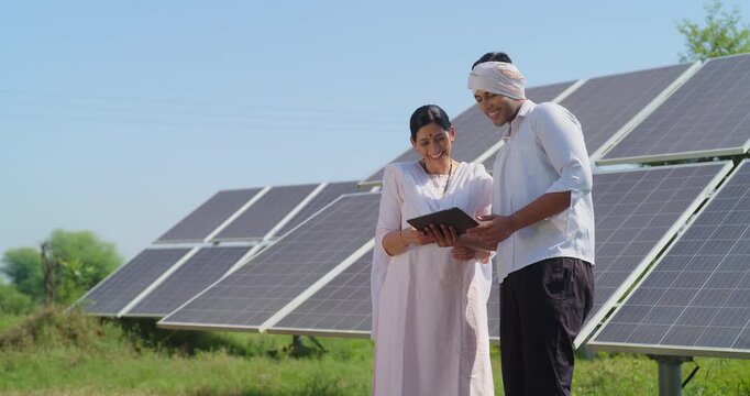 Indian mature adult woman hold tab look screen visit farm field do work check record data at rural area ground place female talk speak share eco friendly solar panel power plan idea to young male