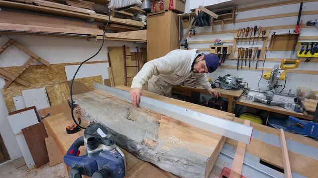Authentic DIY Hobbyist Checking Flatness of Large Live Edge Wood Slab with Metal Straightedge in Messy Home Garage Workshop Wide Angle Handheld Tracking Shot