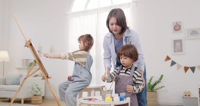 Single mother coaching preschooler son during painting session, daughter paints on easel canvas, quality time with young children, playful learning in bright playroom, early childhood development home
