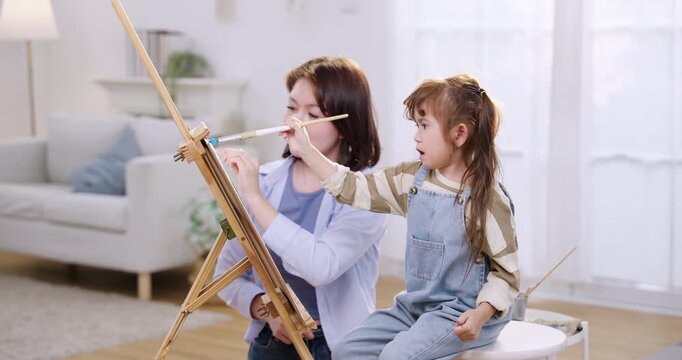 Asian single mother claps encouragement while preschool daughter paints on easel, indoor family lifestyle playtime turns into childhood development lesson, nurturing care, sibling art studio nearby