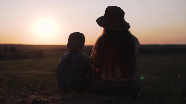 Mother and child sit watching sunset in field wearing caps. Woman with hat sits with boy outdoors at sunset. Family watches golden hour together. Mother and boy sitting in field at sunset.