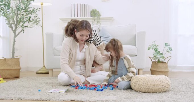 Asian single mother coaches son stacking blocks while daughter observes, learning together on rug in living room. Gentle parenting supports milestone, homeschooling preschool game, family bonding.