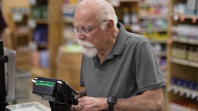 Senior man scans his groceries and pays at the self checkout in a grocery store during a busy shopping hour.