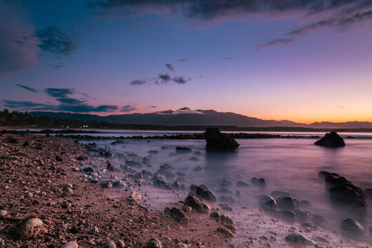 View of the serene shore with rocks meeting the tranquil sea under a twilight sky, painting the scene in shades of purple and orange, Mati, Davao Region, Philippines.