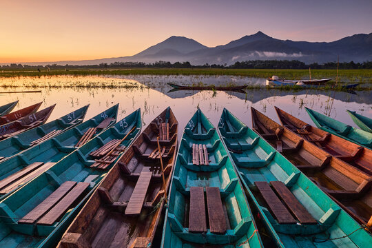 View of vivid teal boats lined up on the still lake, reflecting the distant mountains under the pastel dawn sky, Semarang Regency, Central Java, Indonesia.