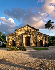 Fototapeta premium An aged, weathered church sits under a dramatic sky. Its facade is detailed, and a tall palm tree rises nearby. The light casts soft hues