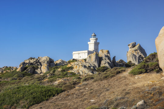View of a white lighthouse standing tall atop rugged rocks, surrounded by dry grass and green shrubs under a clear blue sky, Capo Testa, Sardegna, Italy.