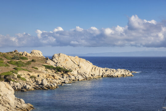 View of rugged, sun-kissed granite rocks meet the turquoise sea under a vast blue sky dotted with fluffy white clouds, Capo Testa, Sardegna, Italy.
