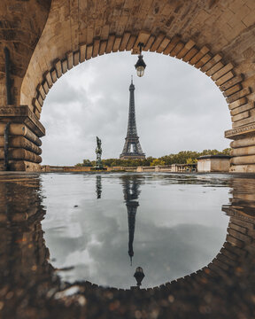 view of the Eiffel Tower piercing the grey sky, reflected in the still waters, framed by ancient stone arches, Paris, France.
