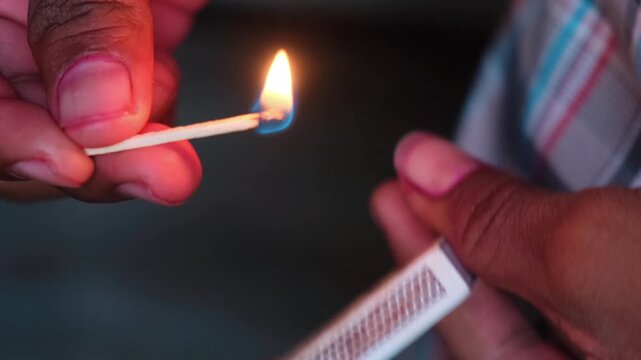 Close-up of a hand striking a matchstick against a matchbox with a bright flame and smoke