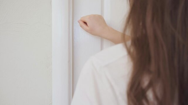 Back view of woman knocking with fist on white wooden door concept of waiting asking for entry and everyday communication