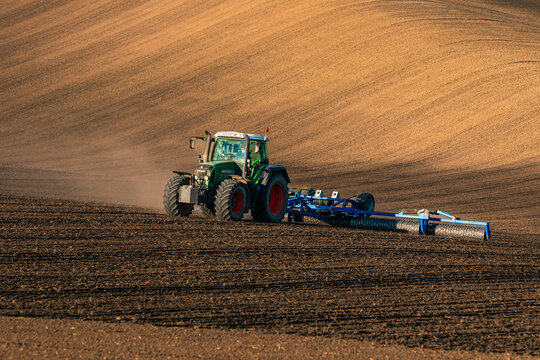 View of a green tractor rolls across the dark, freshly tilled earth, contrasting with the golden, undulating hills in the background, Sardice, South Moravian Region, Czechia.