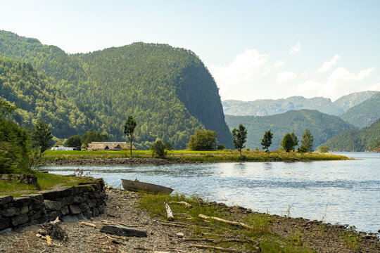 View of shimmering waters meet verdant hills, punctuated by a rustic boat resting on the rocky shore, under a vast sky, Bolstadoyri, Vestland, Norway.