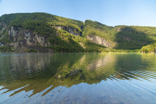 View of the serene waters reflecting the majestic, verdant mountains under a bright sky, creating a harmonious blend of nature's beauty, Bolstadoyri, Vestland, Norway.