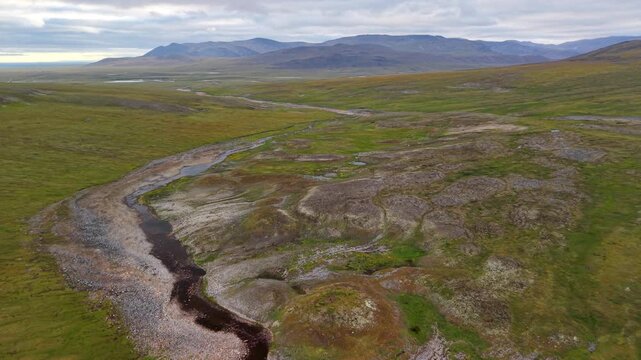 Aerial view of braided river moving through foothills behind Lavrentiya. Gravel bars and oxbow traces draw geometry across tundra. Overcast polar summer light keeps color and detail balanced