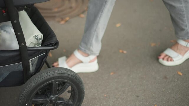 Caucasian feet in sandals pushing stroller wheel on asphalt, closeup detail of tire, basket storage and jeans hem, everyday parenting routine and practical care moment tied to lifestyle decree