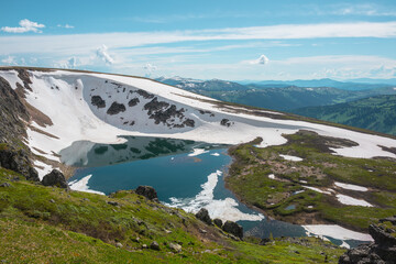 Scenic sunlit landscape with alpine lake in rocky snowy cirque near stone hill top during thaw. Ice floats in mountain lake among rocks and snows with view to forest mountain range under cloudy sky. © Daniil