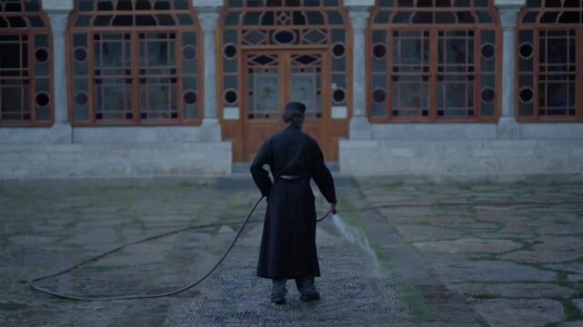 Solemn man in a black cassock spraying water from a hose in a stone courtyard in Greece