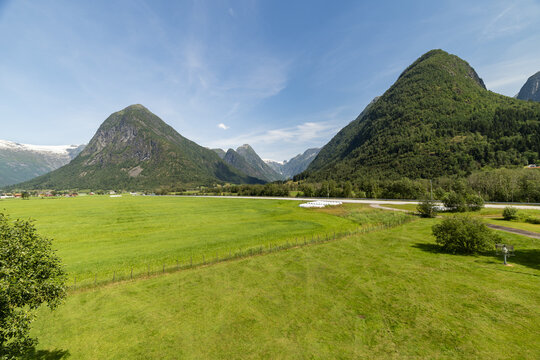 View of the vibrant green field stretches toward the base of towering mountains under a clear blue sky, a serene summer landscape, Fjaerland, Vestland, Norway.