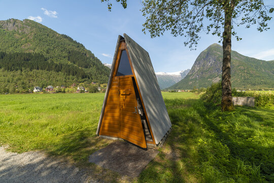 View of a triangular wooden structure nestled in a vibrant green field, framed by majestic mountains under a clear blue sky, Fjaerland, Norway.